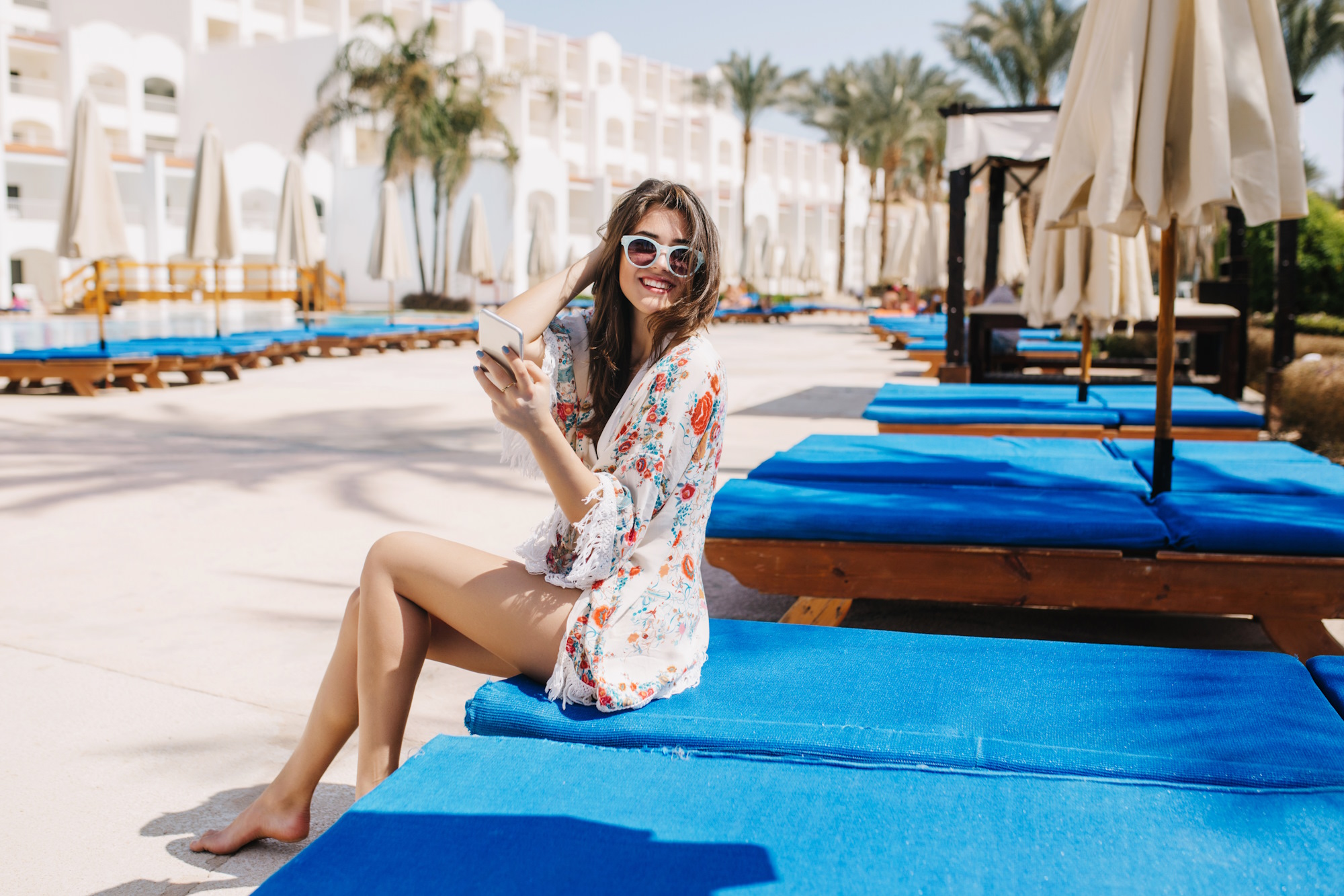 Joyful barefooted lovely girl resting on blue chaise-longue and smiling to camera with palm trees on background. Portrait of cheerful young brunette woman sitting on trestle-bed with phone